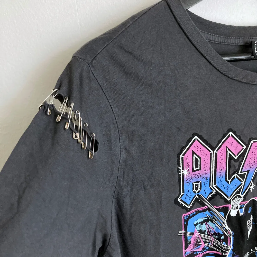 AC/DC t-shirt med säkerhetsnålar i följsamt material. Mått i cm: bröstvidd 50; axelbredd 41; längd fram från axeln 61; ärmlängd 22.. Skjortor.