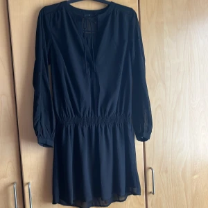 Långärmad klänning i lätt tyg - Svart skir tyg, långärmad, resår i midjan. Knytband och knapp på framsidan av överdelen strl 38 generös i storlek, luftig tyg