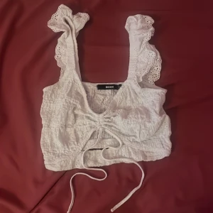 Vit spetsblus från Bik Bok - En vit spetsblus från Bik Bok med volangdetaljer på axelbanden och knytning framtill. Blusen är ärmlös och har en croppad passform.