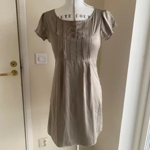Klänning stl S - Söt och enkel klänning men med fina snitt och detaljer. Kommer från Vila. Fint begagnat skick. Dragkedja i sidan. Knappar fram, endast för detaljer. En fin grå nästan metallic färg.  Har använts till dop men passar bra till sommarfest, bröllop tex. 