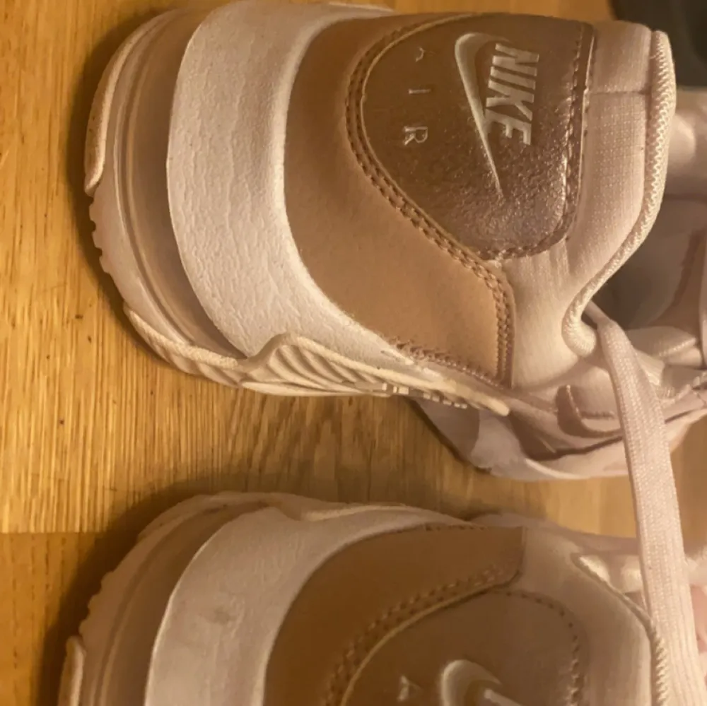 Ett par Nike Air Max sneakers i beige,vit och rosa färg. Skorna har en synlig luftdämpning i sulan och är dekorerade med Nike-logotypen på sidan och hälen. De har snörning framtill och är tillverkade i syntetmaterial.. Skor.