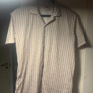 Beige och vit randig skjorta - En beige och vit randig skjorta med korta ärmar. Skjortan har en klassisk krage och knappar framtill. 