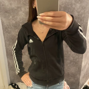 Adidas luvtröja med dragkedja - En svart adidas luvtröja med dragkedja, med 3 vita ränder på vardera ärm på sidorna av armarna. Den har slitningar på ena armbågen och på handlederna men som man inte tänker på. Storleken passar också på XS. 