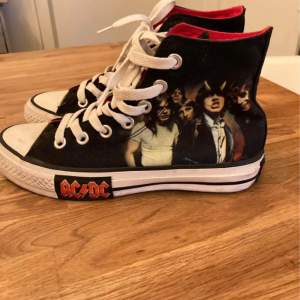 Ett par svarta Converse All Star sneakers med högt skaft och vita snören. Skorna har ett tryck av bandet AC/DC på sidan och texten 'AC/DC' på sulan. Insidan av skorna är röd.