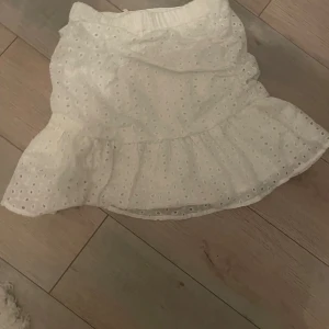 Vit kjol med volangdetaljer - En vit kortkjol med volangdetaljer och broderade hålmönster. Kjolen har en elastisk midja för bekväm passform.