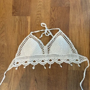 Vit virkad bikiniöverdel - Ett vit virkad strandplagg med knytband i nacken och ryggen som är i storlek S. Överdelen har en dekorativ kant med små hängande detaljer.