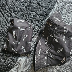 Svart hatt från Moschino - En svart hatt från Moschino med vitt tryck av varumärkets namn. Hatten är tillverkad av 100% polyester och har en bred kant. Den är designad för att vara bekväm och stilren. Som ny!