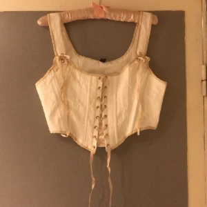 Livstycke, korsett str 42, L - Oanvänt i cremevitt med upphöjda blommor i tyget, beiga satinerande, går reglera mått med band mitt på, förstärkt med inbyggda byglar 2 vardera sida fram, fodrad. Omkr bredaste delen armhåla 98-102 cm, omkr  midja 86-90 cm, fr axel ner fram 50 cm 