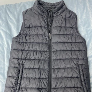Svart dunväst från Everest - En svart dunväst från Everest med dragkedja framtill. Västen är quiltad och har en fin krage som kan vikas värme. Perfekt för våren, sommar och höst. en härlig väst som är enkel att styla!