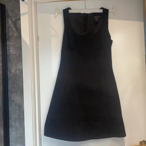 Svart ärmlös klänning från Mini Suit - En elegant svart ärmlös klänning från Mini Suit. Klänningen har en enkel design med knappar på baksidan och en rak passform. Perfekt för formella tillfällen eller som en stilren vardagsklänning.