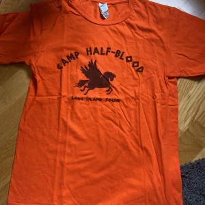 Camp Half-Blood T-shirt  - Orange t-shirt med tryck från böckerna om Percy Jackson. Fint skick, endast använd ett fåtal gånger. En mycket liten fläck på tröjans framsida men annars inga skador. Storlek S. Skriv vid funderingar😊