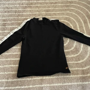 Svart tröja  - svart långärmad tröja med en vit rand som löper längs med ena ärmen, där det också finns vita knappar.