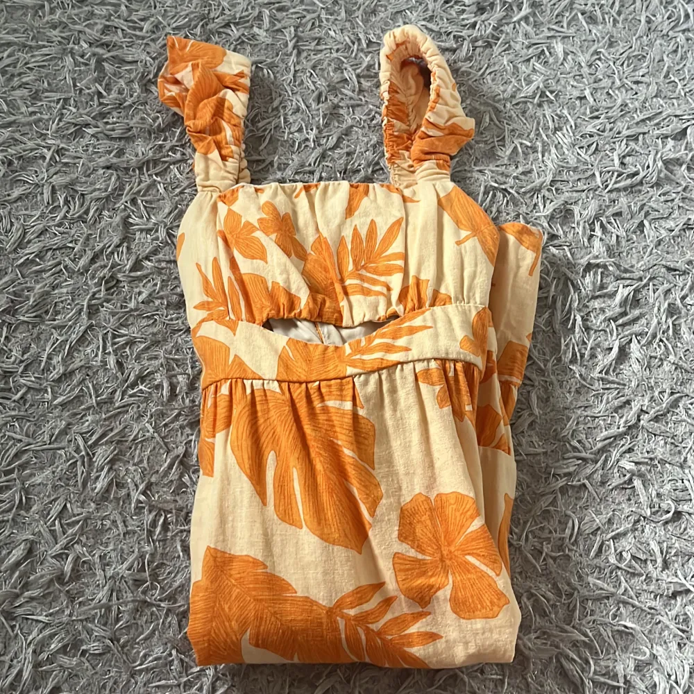 En somrig klänning från Zara med ett blommigt mönster i orange och beige. Klänningen har breda axelband med volangdetaljer och en smockad överdel. Den är tillverkad i ett lätt och bekvämt material, perfekt för varma dagar.. Mekot.