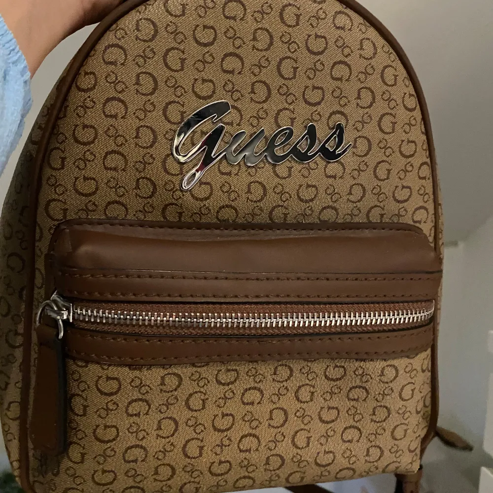 Säljer en snygg brun ryggsäck från Guess. Väskan har ett mönster med Guess-loggan över hela ytan och en stor Guess-logga i silver på framsidan. Perfekt för vardagen! Helt oanvänd i nytt skick. Köpte den på rea för 800kr. Pris går att diskutera . Väskor.