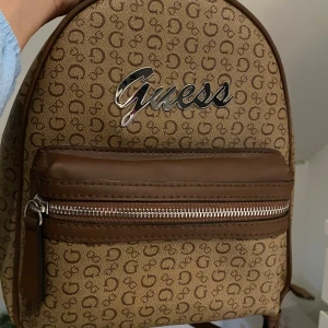 Brun ryggsäck från Guess - Säljer en snygg brun ryggsäck från Guess. Väskan har ett mönster med Guess-loggan över hela ytan och en stor Guess-logga i silver på framsidan. Perfekt för vardagen! Helt oanvänd i nytt skick. Köpte den på rea för 800kr. Pris går att diskutera 