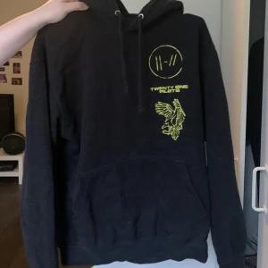 Svart hoodie från Twenty One Pilots - Säljer en svart hoodie från Twenty One Pilots med gult tryck. Den har en stor ficka fram och en luva med dragsko. På framsidan finns bandets logga och en örn, och på baksidan står det 'Bandito' med en stor örn. Perfekt för fans av bandet! 🖤💛 Längst ner står också Line up-en för hela albumet trench. Tröjan är ursprungligen från EMP och är i väldigt bra skick. 