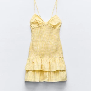 Gul rutig klänning med volangdetaljer - Säljer en superfin gul rutig klänning med volangdetaljer. Klänningen är kort och har smala axelband. Den är perfekt för sommaren och har en söt rosett framtill. Materialet känns lätt och luftigt, vilket gör den idealisk för varma dagar på stranden eller sommarfester. Passformen är tight och klänningen har en härlig, lekfull stil.