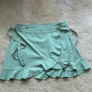 Mintgrön omlottkjol med volang från Anzi Felter - Säljer en superfin mintgrön omlottkjol. Kjolen har en söt volangdetalj längst ner och knytband i midjan. Perfekt för sommaren eller en festlig tillställning. Den är i nyskick och aldrig använd. Passar perfekt till en avslappnad men ändå chic look!