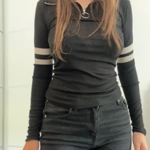 Svart långärmad tröja med dragkedja och vita ränder - Säljer en svart långärmad tröja i storlek S. Tröjan har en dragkedja med en ringdetalj vid halsen och vita ränder på ärmarna. Den är tight och passar perfekt för höst och vinter. Materialet känns som bomull och är väldigt bekvämt. Perfekt för både vardag och fest!