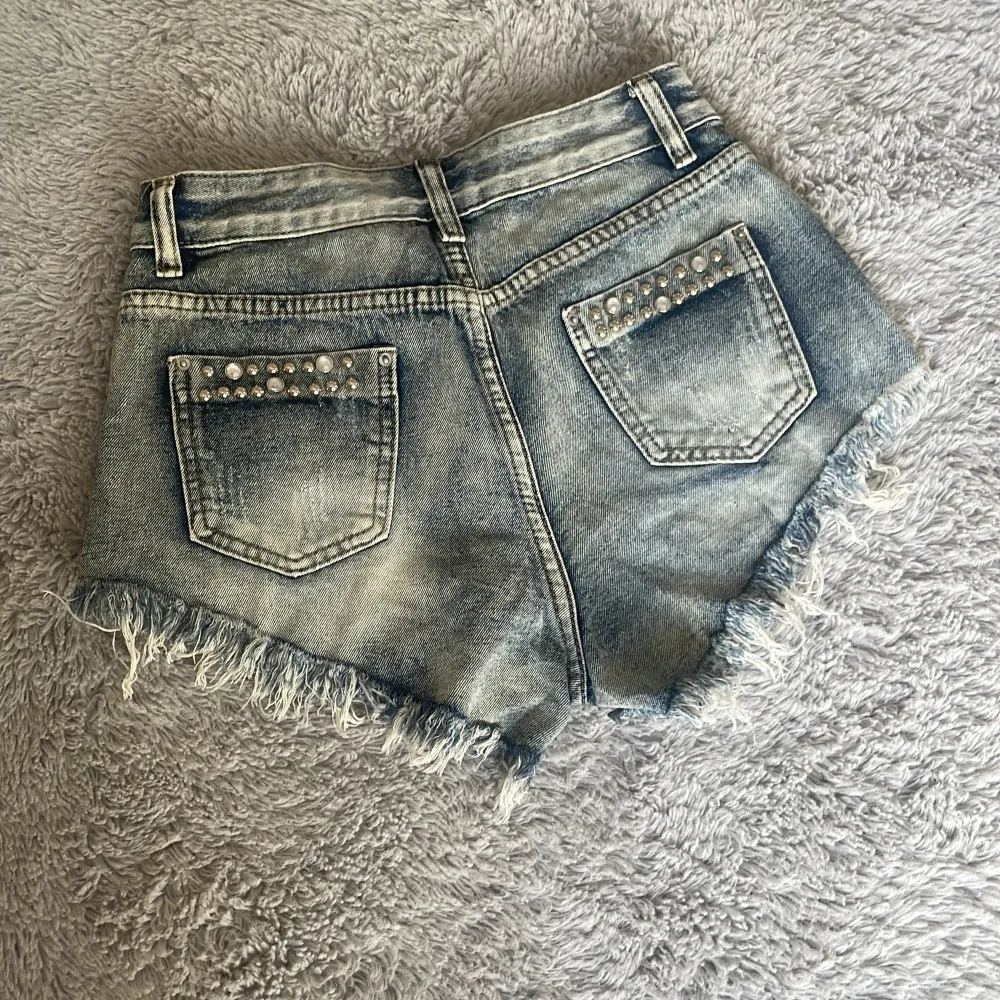 Säljer ett par snygga helt oanvända jeansshorts i storlek XS. De är ljusblå med coola slitningar och pärldetaljer både fram och bak. Shortsen har fransiga kanter och en låg midja. Perfekta för sommaren och festivaler! 🩷👌. Shorts.