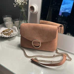 Beige mocka axelväska med kedja - Säljer en superfin gammalrosa axelväska i mocka. Väskan har en elegant design med en stor guldring på framsidan och en kedja som axelrem. köpte för 2 år sedan men har knappt använt dem💕