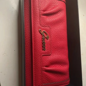 Röd plånbok från Guess - En elegant röd plånbok från Guess med texturerad yta. Plånboken har ett inre fack med dragkedja och är dekorerad med Guess-logotypen på framsidan.