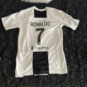 Vit och svart fotbollströja med Ronaldo 7 - Säljer en vit och svart fotbollströja från Juventus med Ronaldo 7 på ryggen. Tröjan är i nyskick och har korta ärmar. Perfekt för alla fotbollsfans som vill visa sitt stöd för sitt favoritlag och spelare!