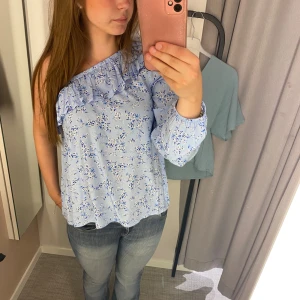 Blå blommig offshoulder-topp från Lindex - Säljer en superfin blå offshoulder-topp från Lindex med blommigt mönster. Toppen har en volangdetalj längs halsen och är perfekt för sommaren. Den är i storlek 164 och passar både till vardags och fest. Toppen är i nyskick, använd en gång!