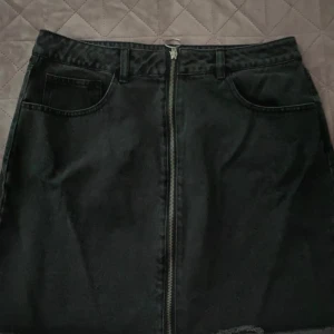 Svart jeanskjol med dragkedja - En svart jeanskjol med dragkedja framtill. Kjolen har två framfickor och bälteshällor. Helt ny saknar prislapp dock.