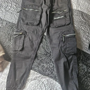 Svarta cargobyxor från Black Squad - Svarta cargobyxor från Black Squad med flera fickor och dragkedjor. Byxorna har en elastisk midja och är tillverkade i Pakistan.