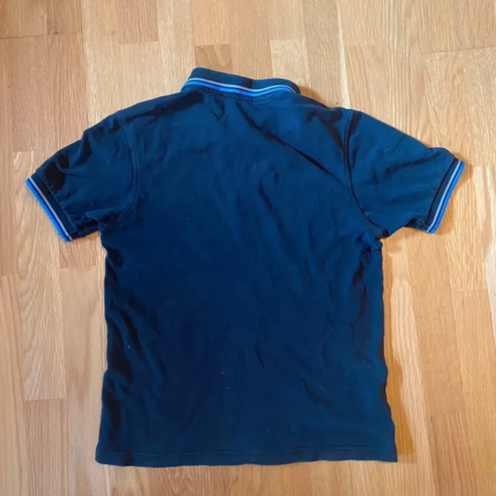 Svart Fred Perry tröja med blåa och grå sträck hade hål fast har lagat är annars i bra skick. T-shirts.