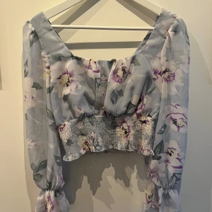 Blommig croppad blus - En blommig croppad blus med långa ärmar och elastisk midja. Blusen har ett mönster av vita och lila blommor på en ljusblå bakgrund.
