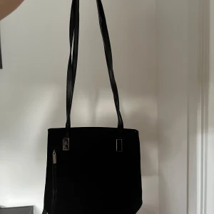 Svart axelväska - En svart axelväska med dragkedja. Väskan har en enkel och stilren design med en lång axelrem. Insidan av väskan har flera fack med dragkedja för extra förvaring.