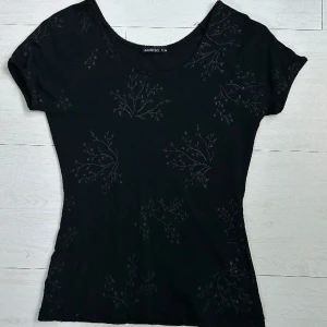 Svart T-shirt med mönster - Svart T-shirt med glittrigt blommigt mönster! :)  Figursydd T-shirt som jag köpt secondhand men aldrig kommit till användning tyvärr! Materialet är lite halvgenomskinligt som visas på bild 2🥰