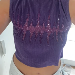 Lila topp - Lila topp med mesh detaljer över bysten. Med pärlor som fint broderats på. Polo/ högre krage med en dragkedja där bak. Tyget är glittrigt. Finns ingen lapp men passar mig bra som är en stl S, den har lite stretch i sig så kan passa större storlekar