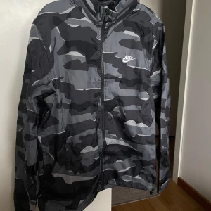 Nike Camo Windbreaker storlek M - Grå / svart vind jacka från nike i storlek M, har en svart luva i kragen som kan vikas in/ut (se bilder)