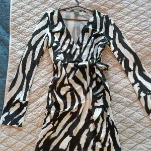 En zebra färgad omlottklänning i storlek S från Gina Tricot i helt nytt och oanvänt skick! Klänningen har en slits vid båda ärmarna och knytning vid ena sidan av midjan. 