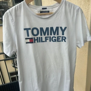 Tommy Hilfiger Organic Cotton Tee, boys size 176cm - Vit tischa från Tommy Hilfiger som är i nästintill nyskick! Killstorlek 176cm men funkar även som herrstorlek X-small/Small. Köpt på Kidsbrandstore för 499kr. Kan mötas i Sthlm!