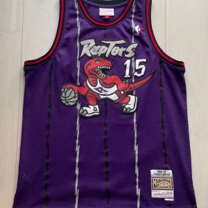 Toronto Raptors Jersey (1998/99)- Mitchell & Ness - Toronto Raptors hemmaställ från NBA-säsongen 1998/99 med Vince Carter på ryggen. Köpt 2022 men är i mycket gott skick då den använts enstaka gånger. 