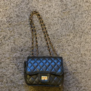  Axelremsväska - Liten axelremsväska i svart med guldfärgade detaljer. Dragkedjan inuti väskan är trasig, i övrigt är väskan i gott skick.