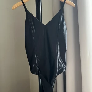 Bodysuit med tunna axelband - V-ringad bodysuit från Bik Bok som sitter som en smäck, glimrande svart tyg och justerbara, tunna axelband. Aldrig använd, lappen sitter kvar.  Kan fraktas eller mötas upp i Göteborg.