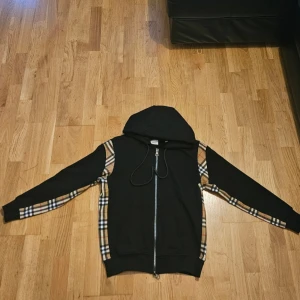 Svart hoodie med rutiga detaljer - Snygg svart hoodie med dragkedja och klassiska beige-rutiga detaljer längs ärmar och sidor. Utrustad med huva och fickor framtill. Perfekt för en trendig och avslappnad stil.