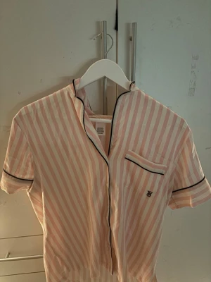 Signature satin short pyjamas - Kortärmad pyjamaströja från Victoria's Secret i ljusrosa och vita ränder. Klassisk krage, svart passpoal och bröstficka med liten logga. Mjukt, glansigt material som känns lyxigt mot huden. Perfekt för mysiga kvällar och chill dagar hemma.