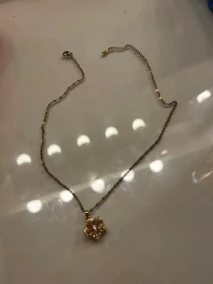 Guldfärgat halsband med blomma - Sött halsband i guldfärg med en blomformad berlock dekorerad med rosa och vita pärlor. Kedjan är tunn och elegant, perfekt för att ge en feminin touch till din outfit. Blomman har detaljerade blad och gnistrar fint i ljuset. Blomman går att snurra på