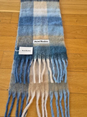 Rutig halsduk Acne Studios blå/beige - Mjuk och fluffig halsduk från Acne Studios i rutigt mönster med nyanser av blått, beige och vitt. Långa fransar i ändarna ger en chill vibe. Perfekt accessoar för att lyfta din outfit med lite färg och stil.