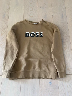 Beige hoodie med BOSS-tryck - Snygg beige hoodie från BOSS med stort svartvitt BOSS-tryck på bröstet. Klassisk modell med rund hals och långa ärmar. Perfekt för en avslappnad och trendig look. Tillverkad i mjukt bomullsmaterial.