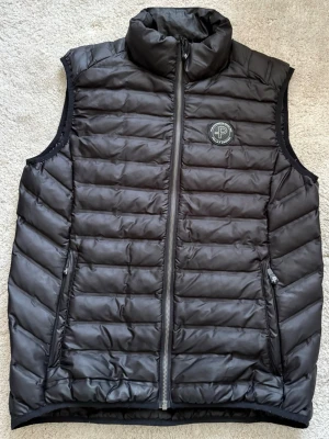 Svart dunväst med ståkrage - Snygg svart dunväst med quiltad design och ståkrage. Västen har två dragkedjeförsedda fickor framtill och en diskret logga på bröstet. Perfekt för lager-på-lager och håller dig varm under kyliga dagar.