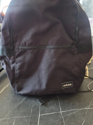 Svart ryggsäck från Adidas - Snygg svart ryggsäck från Adidas med stor vit logga upptill och en mindre logga på framsidan. Ryggsäcken har en stor ytterficka med dragkedja och ett praktiskt bärhandtag. Tillverkad i slitstarkt tygmaterial, perfekt för skolan eller träningen.