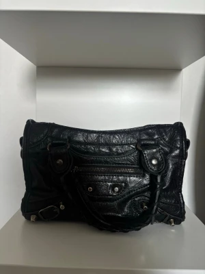 Svart handväska i skinn med nitar - Cool svart handväska i skinn med silverfärgade nitar och dragkedja framtill. Väskan har dubbla handtag och detaljerade sömmar som ger en edgy look. Perfekt för dig som gillar en rockig och trendig stil.