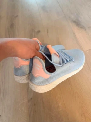  Adidas Pharrell Williams Ljusblå sneakers med rosa detaljer -  Adidas Pharrell Williams Snygga sneakers i ljusblå mesh med rosa detaljer på häl och plös samt vit sula. Skorna har en sportig och modern look med lågt skaft och matchande ljusblå skosnören. Perfekta för dig som gillar stilrena och fräscha färgkombinationer.